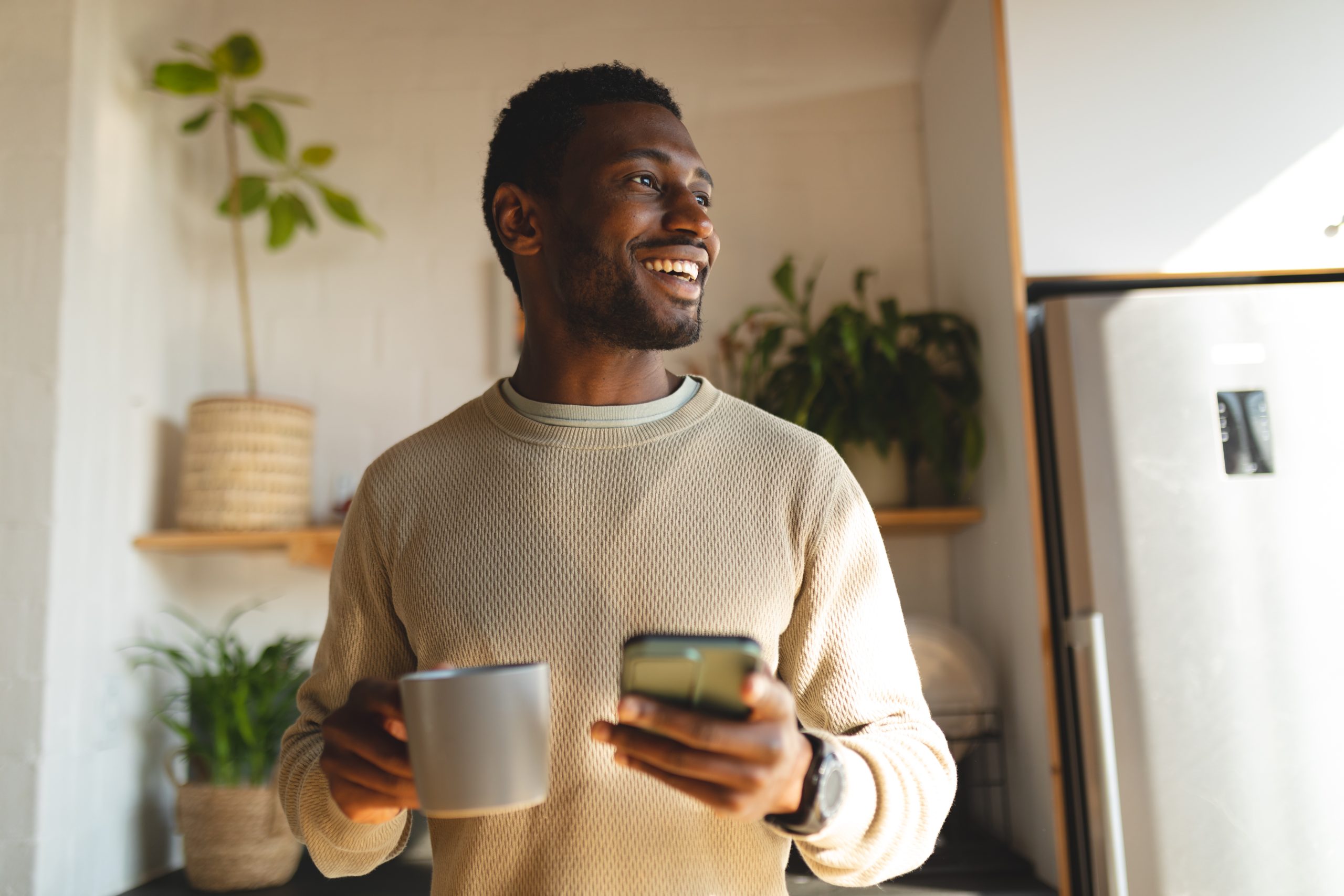 happy african american man using smartphone and drinking coffee in kitchen scaled